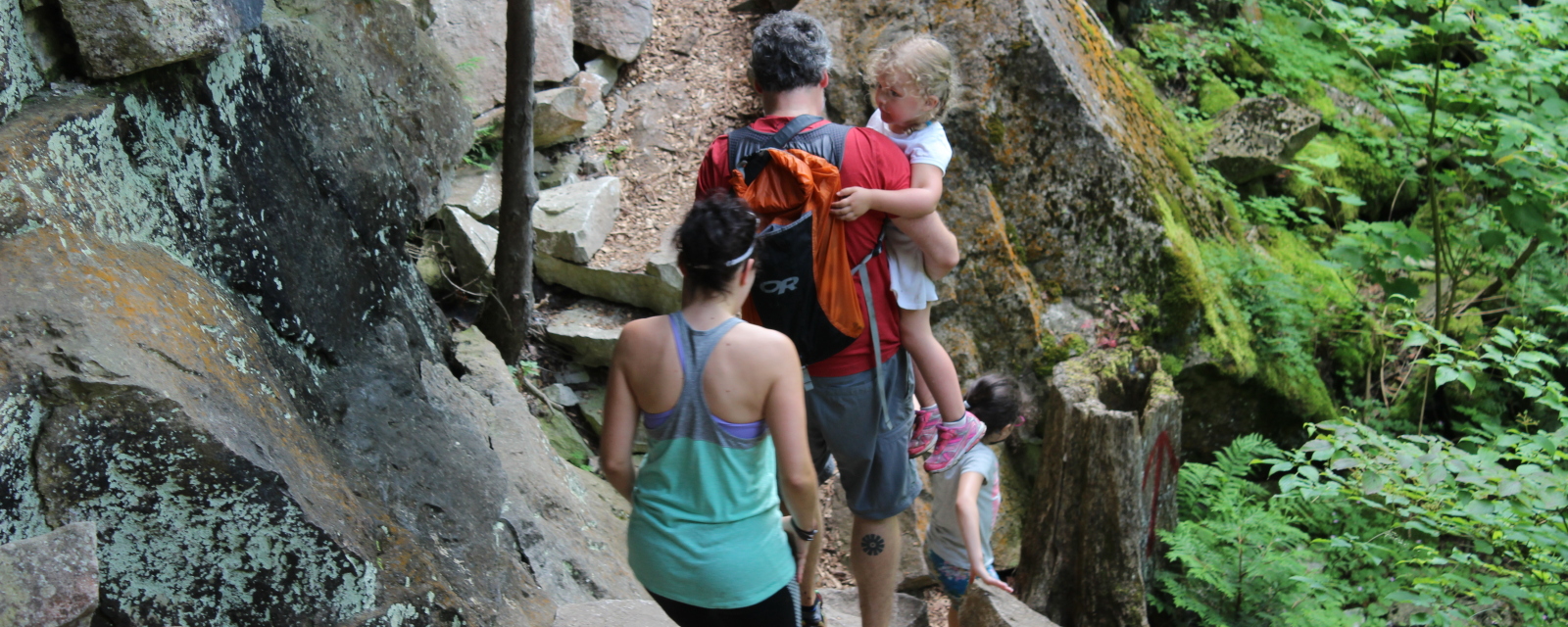 couple hiking on some rocks with their 2 children. Dad is carrying one of the kids