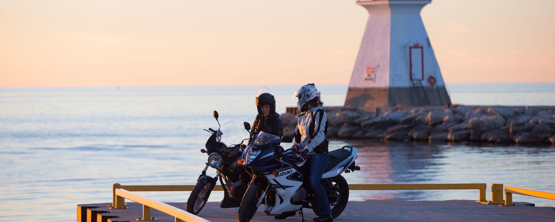 two bikers stopped on a pier, chatting with each other with a lighthouse and the lake in the background.