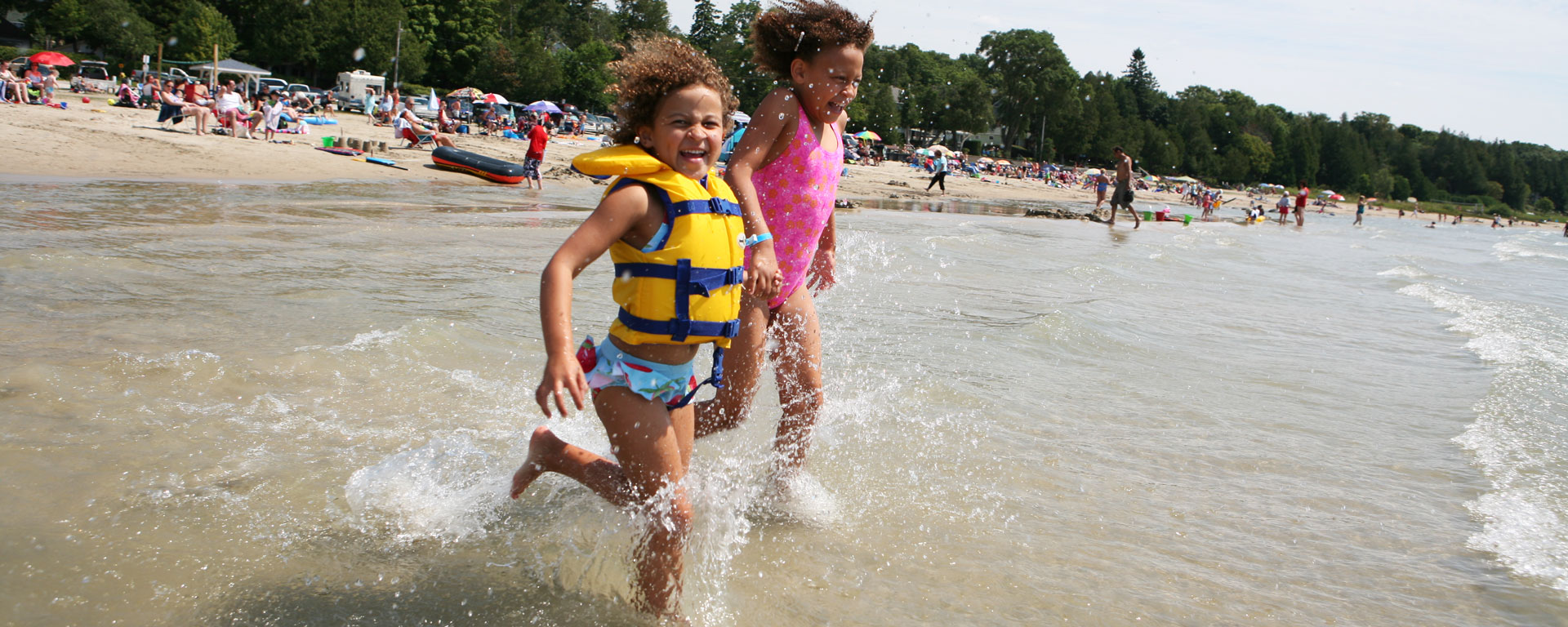 two kids, one in pink the other in a yellow lifejacket splashing and running into the water