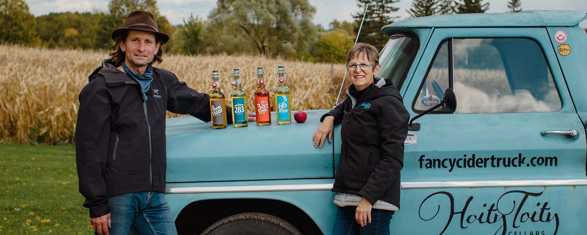 two people leaning against a retro blue truck with ciders on the hood of the truck