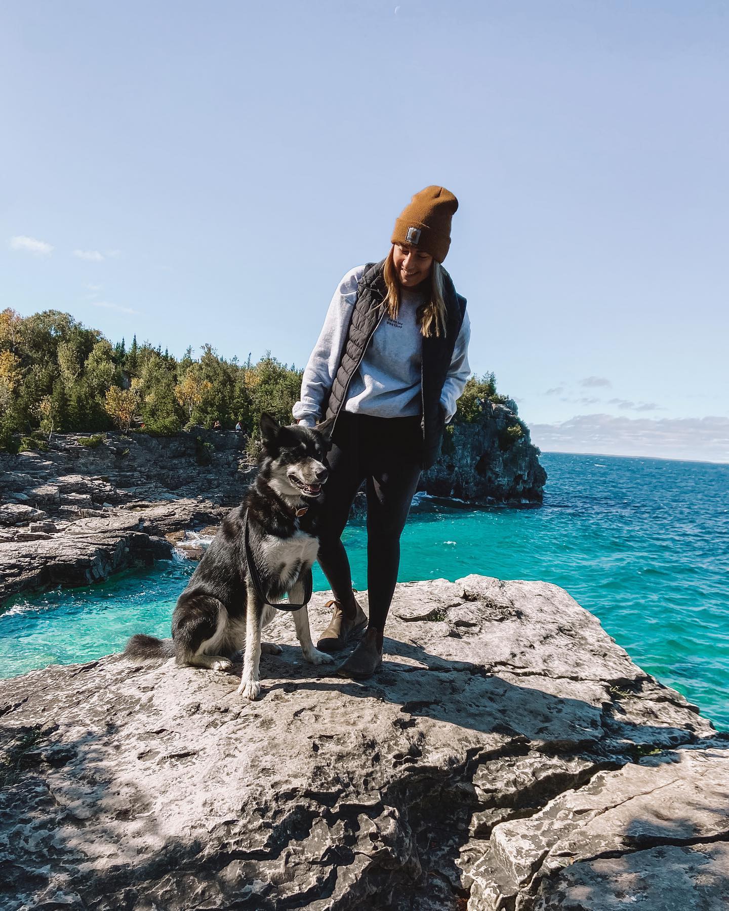 Happy woman posing for the camera with her dog, standing on rocks at Bruce Peninsula National Park with a clear sky and vibrant blue water. photo by @adventuringwithkate