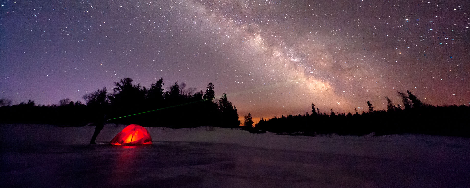 a tent glowing from the light within set on a frozen landscape with a vibrant night sky showcasing the milky way stars