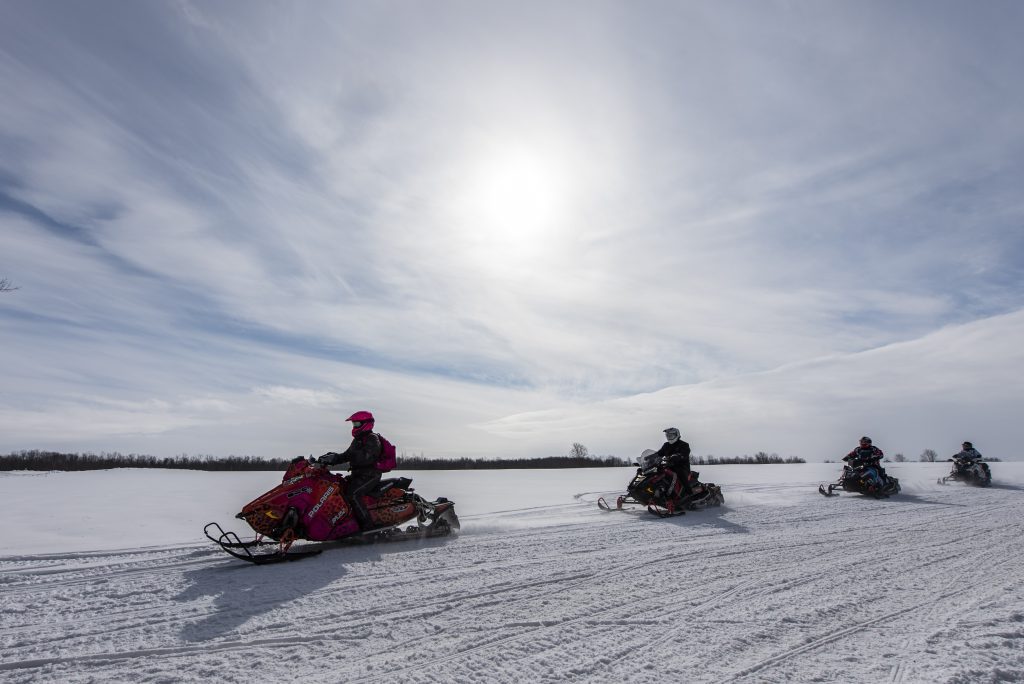 A group of four people riding snowmobiles in an open field with the sun gazing in the blue sky. 